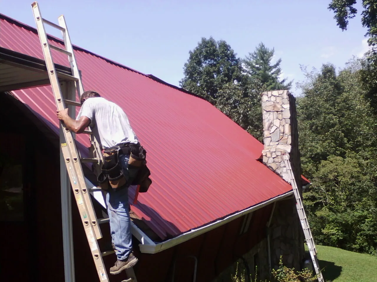 Roofer on a ladder beside a red metal roof and stone chimney — Soffit & Fascia Repair by Roofing Brothers in Redvale